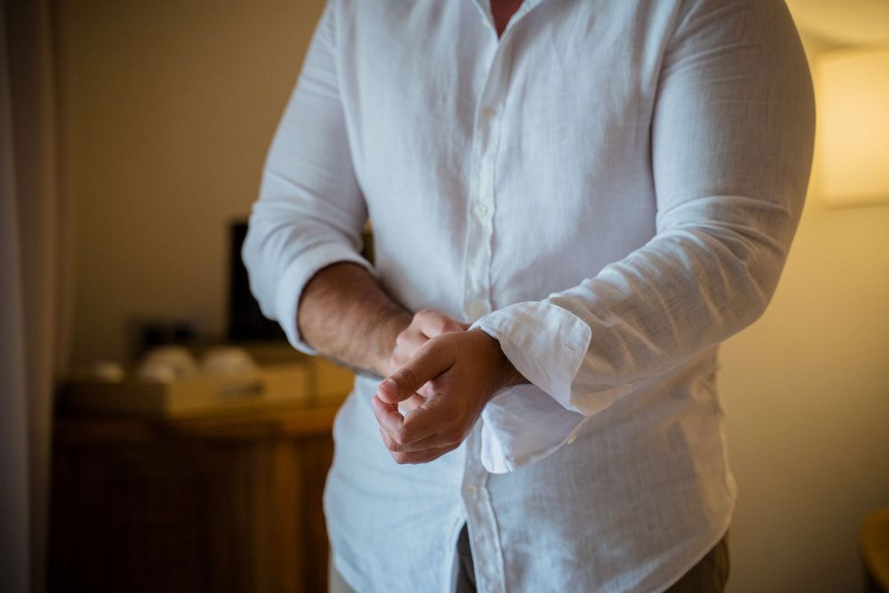 Man in white linen shirt buttoning his cuffs while getting dressed for a formal occasion.