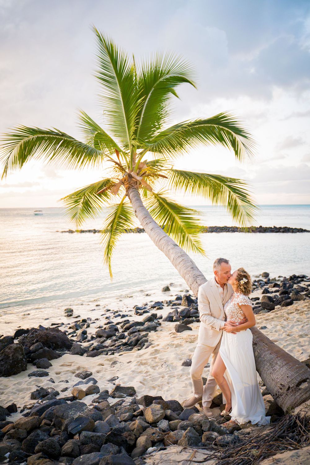Couple kissing under a leaning palm tree on a rocky Hawaiian beach at sunset in wedding attire.