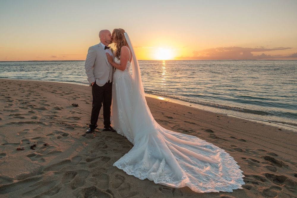 Newlywed couple kissing on a sandy beach at sunset, bride in white gown with long train, groom in gray suit.