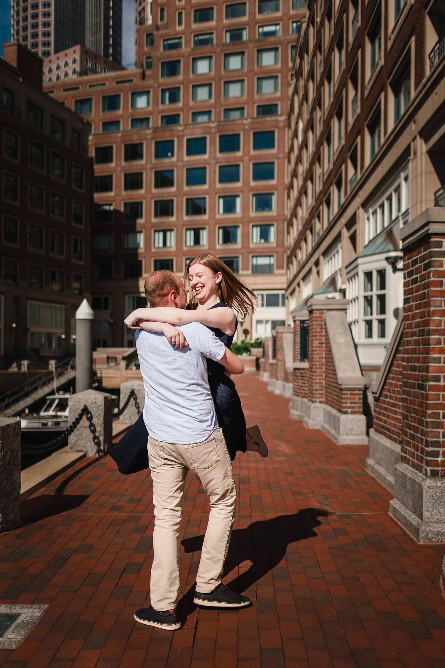 Engaged couple laughing while he spins her around near Rowes Wharf in Boston