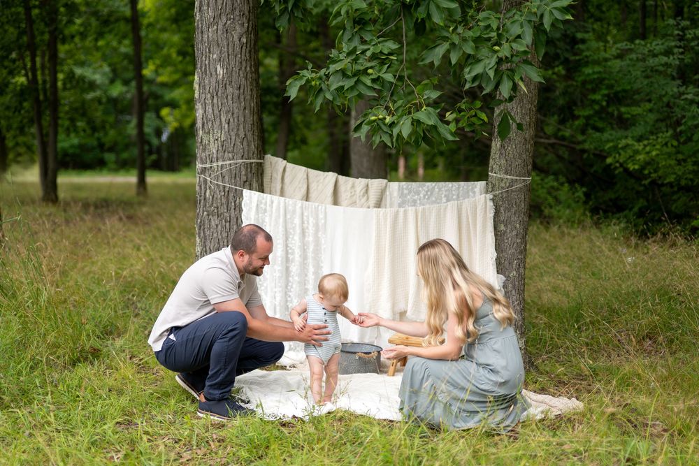Family with baby enjoying outdoor photo session on blanket with lace backdrop between trees in lush green forest.