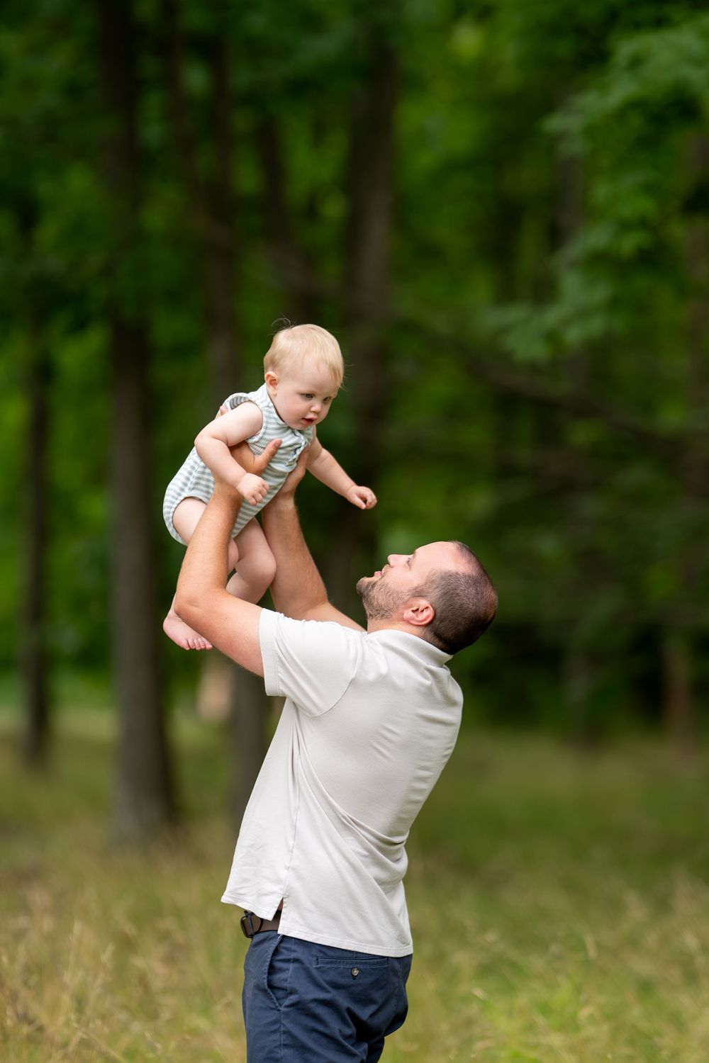 Father joyfully lifting baby in the air outdoors in a green forest park setting.