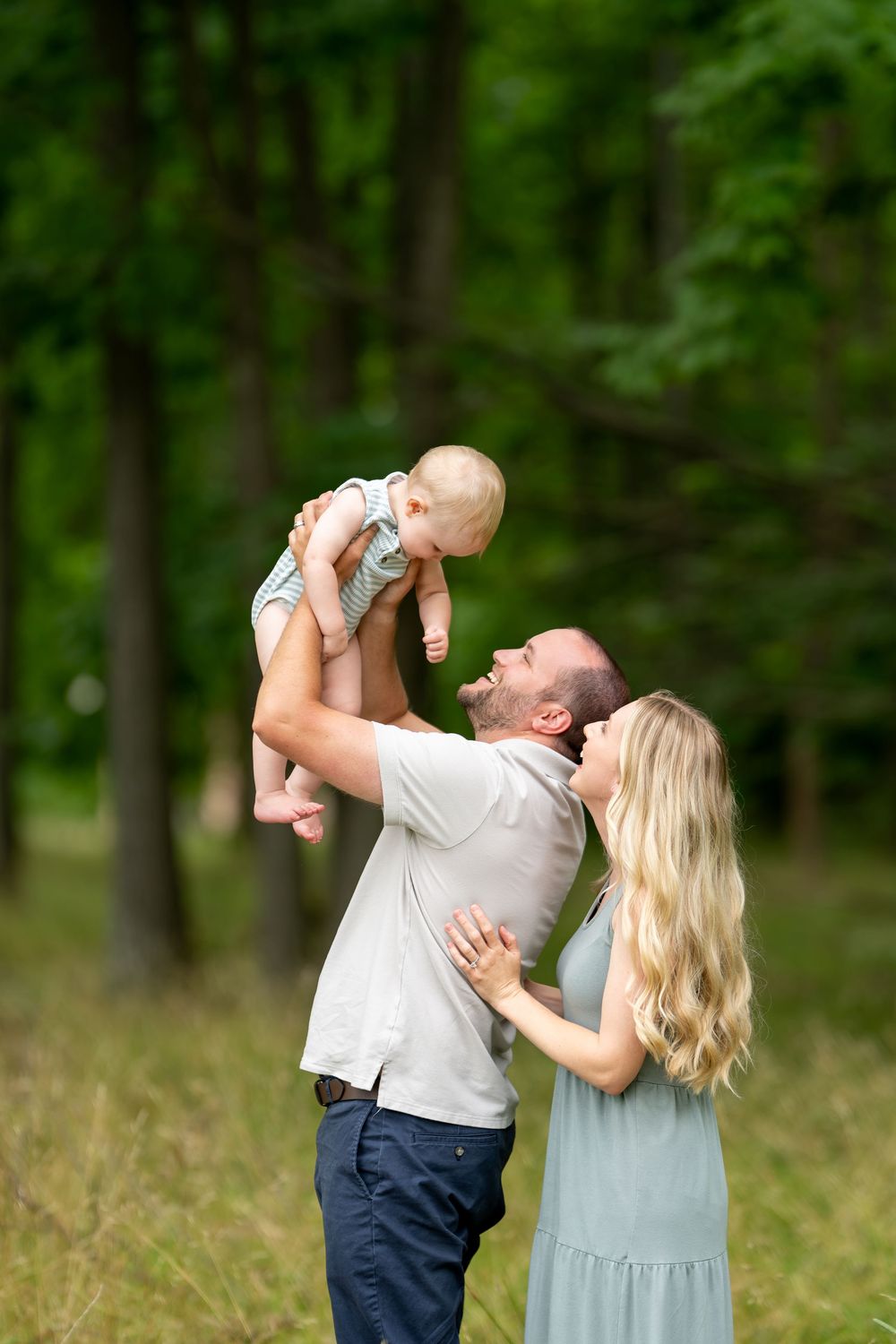 A father lifts a smiling baby overhead while the mother looks on lovingly in a green forest.