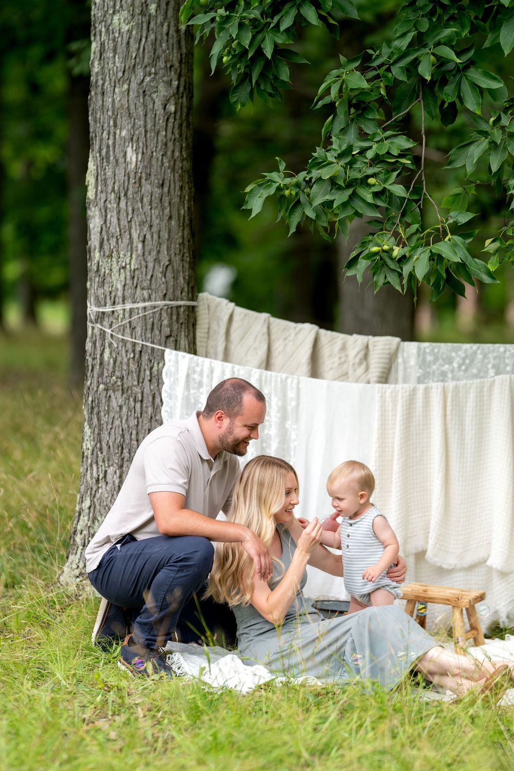 A young family with a baby enjoys an outdoor picnic on grass near hanging white linens and a large tree.