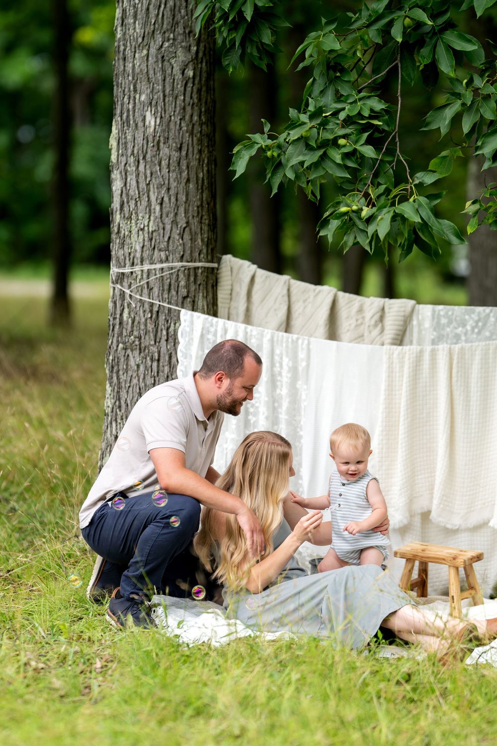 Young family with baby enjoying outdoor picnic in green park with white linens hanging on clothesline.