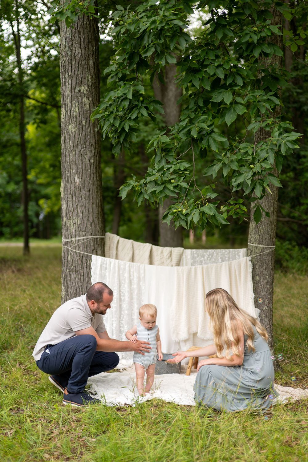 Family with baby enjoying outdoor picnic in green park, with linen backdrop hung between trees.