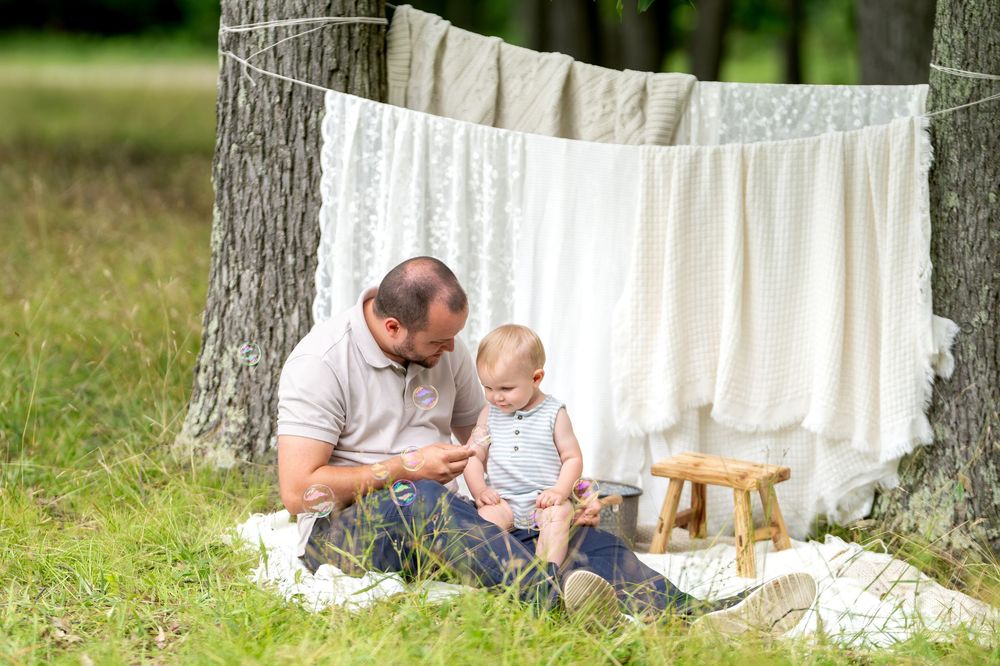 Father sitting on blanket outdoors with baby near laundry line and tree in park setting.