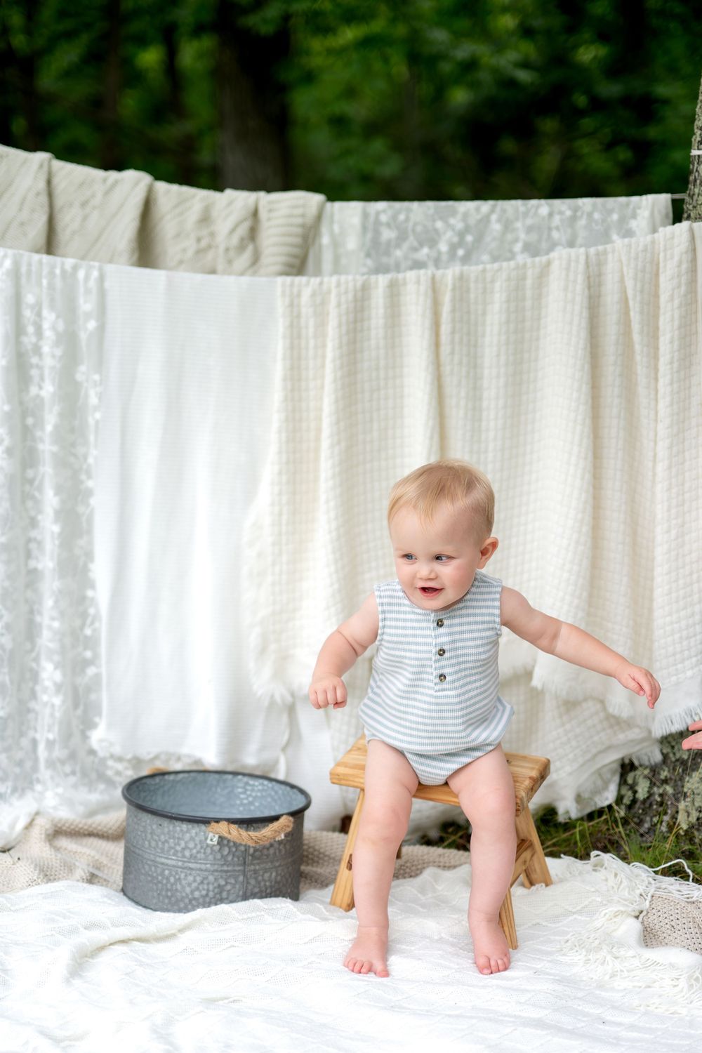 Cute baby in striped romper standing on a wooden stool outdoors with white linens hanging on a clothesline.