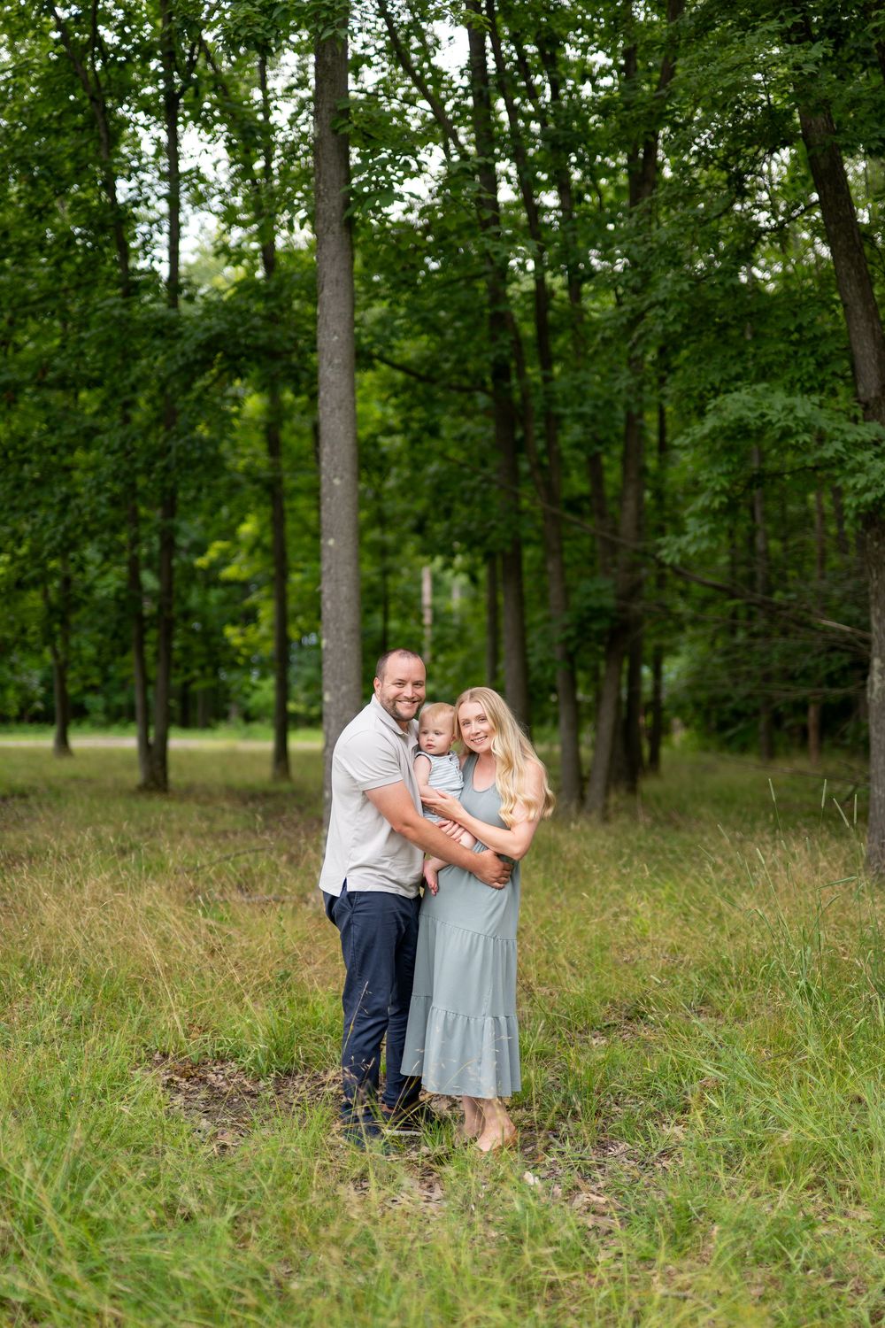 A happy family of three poses in a lush green forest, parents holding their young child in a warm embrace.