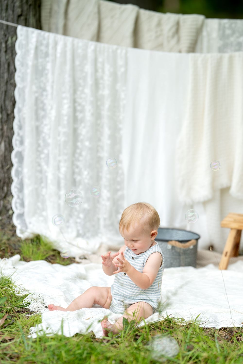 A baby in a striped romper sits on a white blanket outdoors near hanging white laundry and a metal tub.