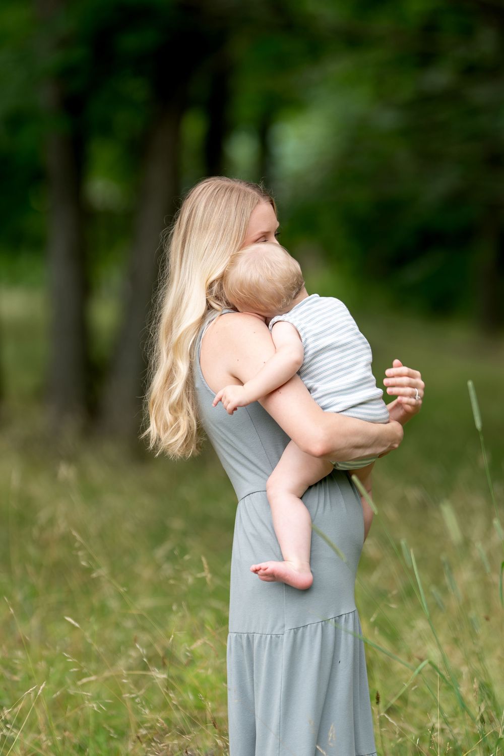 A blonde woman in a gray dress holds a baby outdoors in a lush green field surrounded by trees.