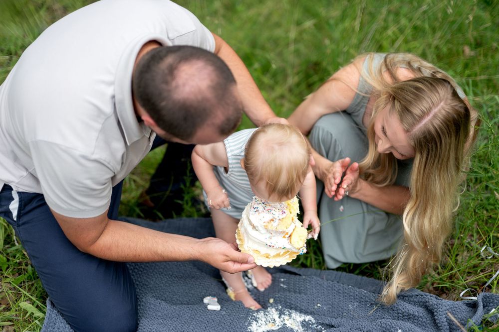 Parents help their baby smash a white cake outdoors on a blanket in the grass.
