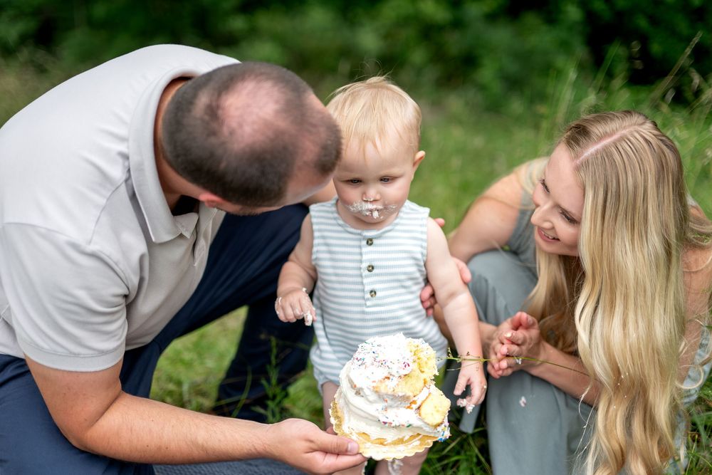 Family celebrating baby's first birthday outdoors, with parents helping toddler smash a white frosted cake.