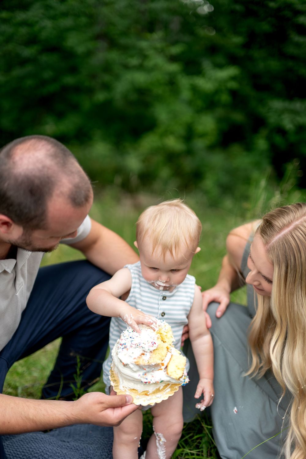 Family celebrates baby's first birthday with a smash cake outdoors in nature.