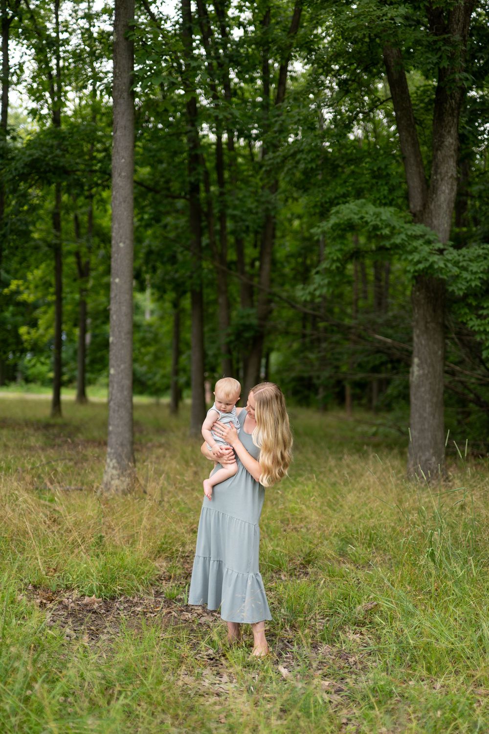 A mother in a sage green dress holds her baby while standing barefoot in a lush green forest clearing.
