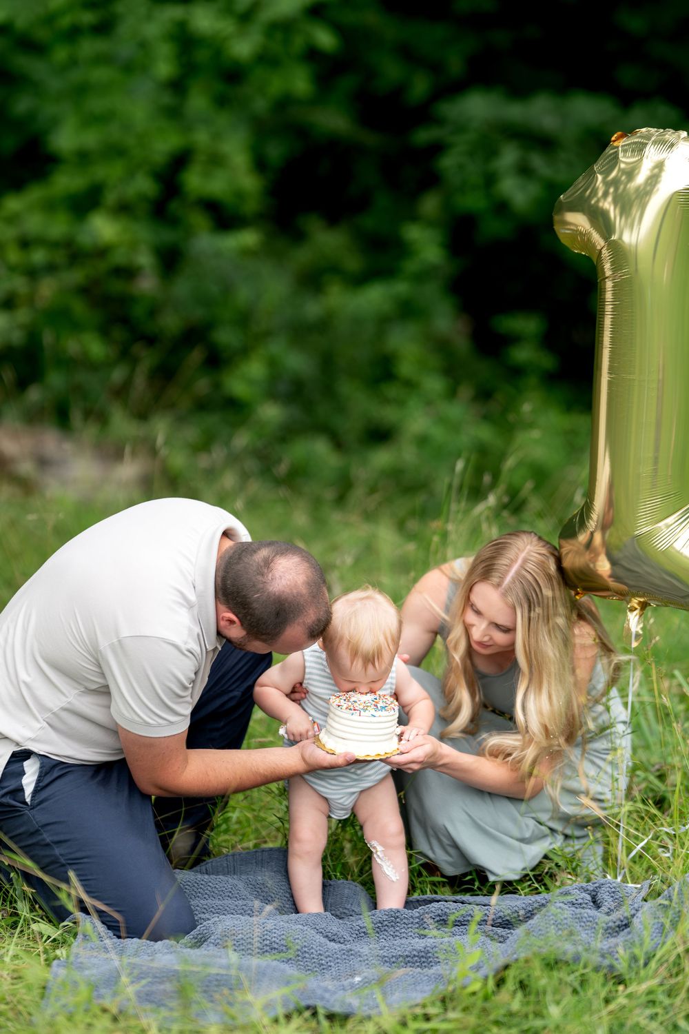 Family celebrating baby's first birthday outdoors with a white cake and gold number one balloon in a green park.