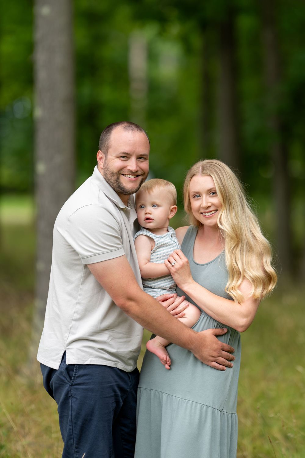 A smiling family of three poses outdoors, with a man and blonde woman holding a baby in a lush green park setting.