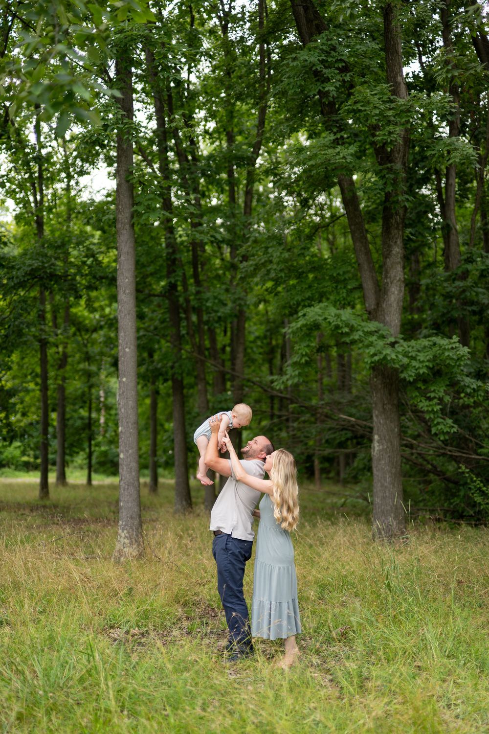 A couple in a green forest clearing lifts their baby joyfully, the mother wearing a sage dress.