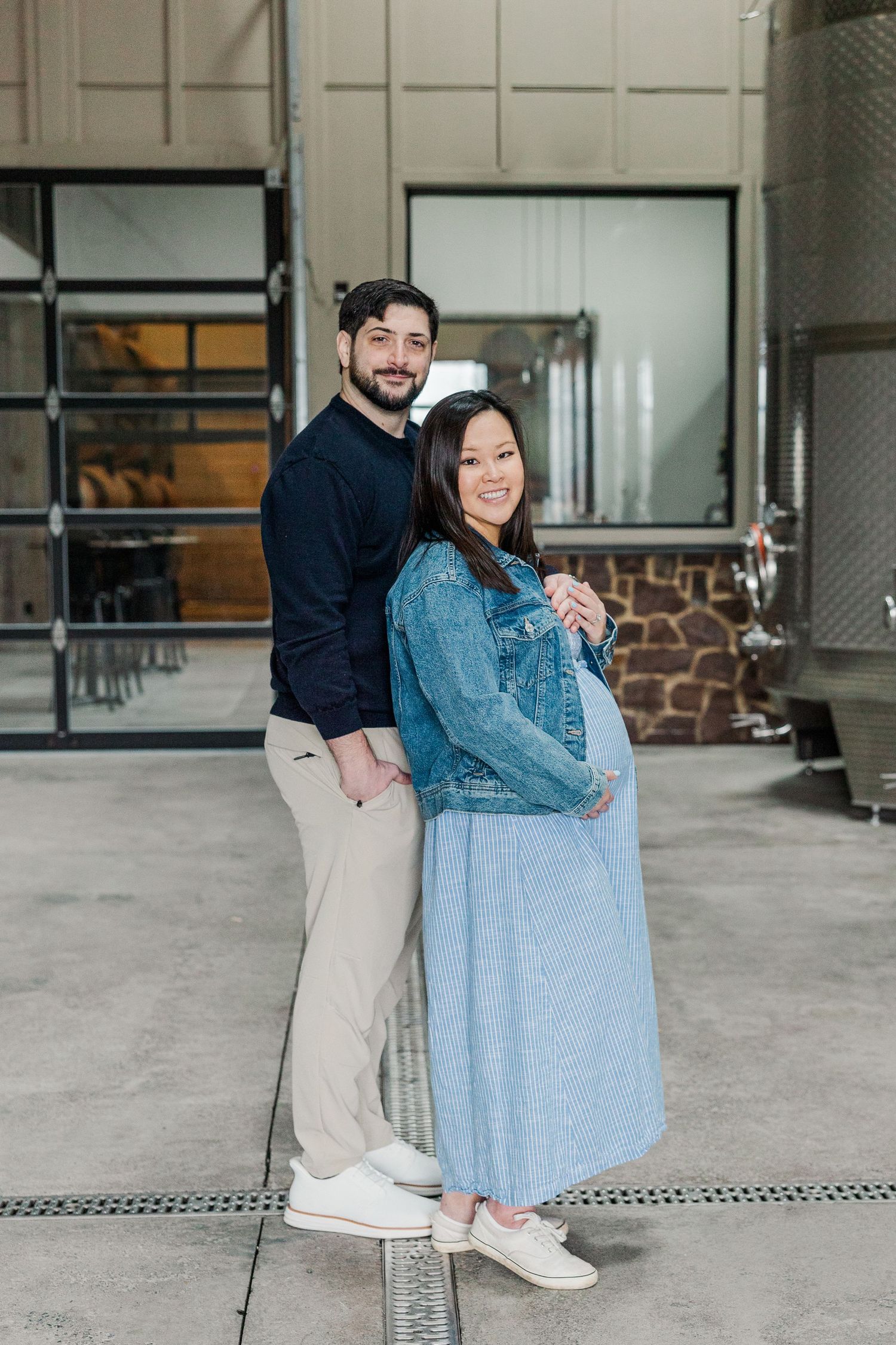 Couple posing for maternity photo shoot, woman in denim jacket and blue dress cradling baby bump, man in navy sweater behind her.