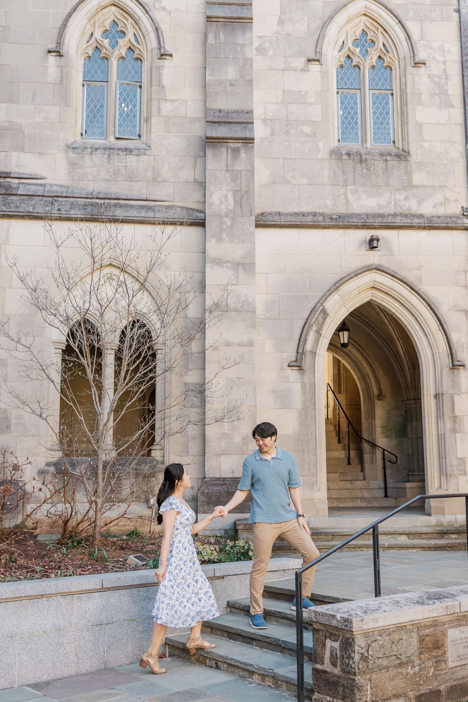 Couple holding hands on stone steps outside a Gothic-style church with arched windows and doorways.