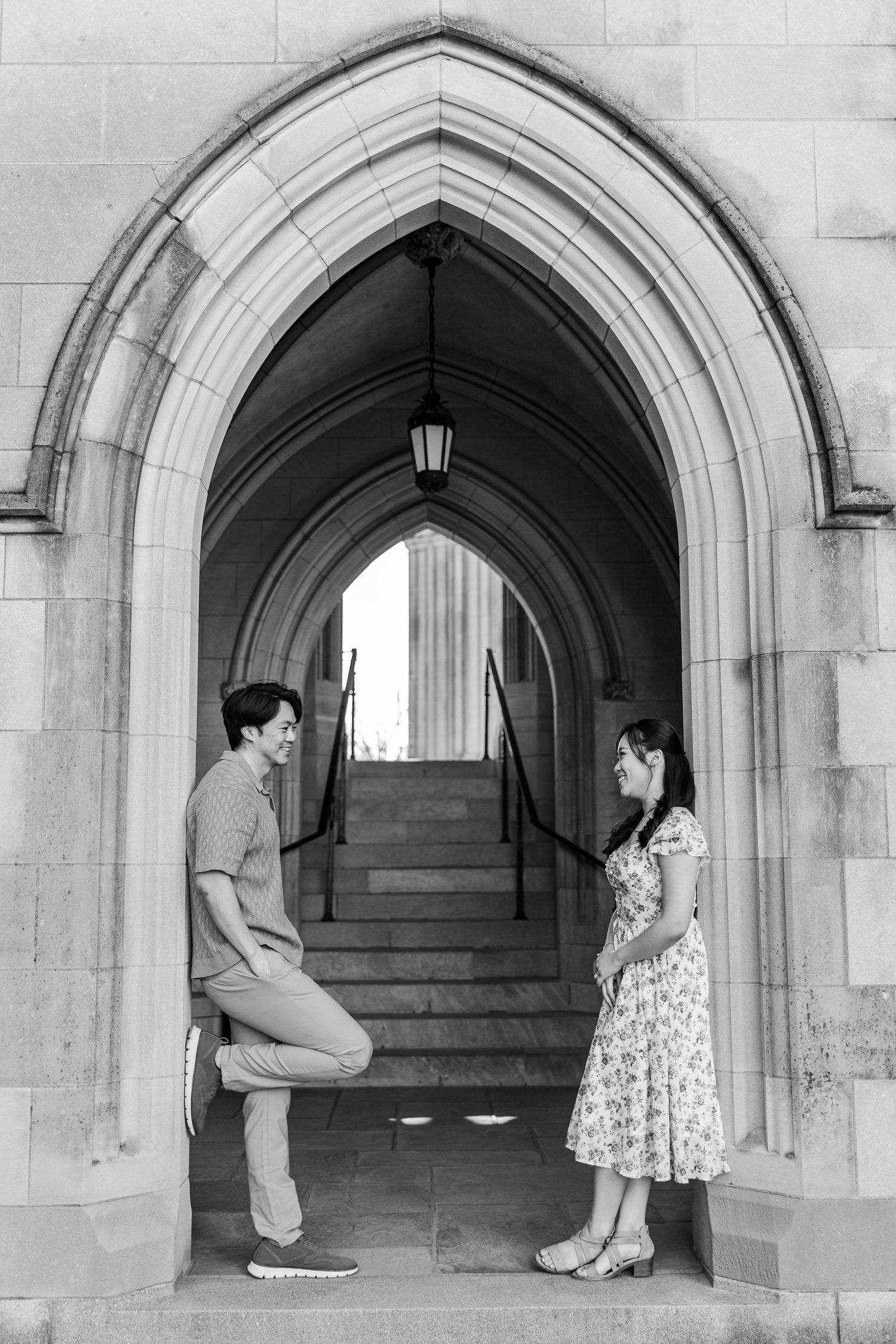 Black and white photo of a couple smiling at each other under a Gothic stone archway with steps behind them.