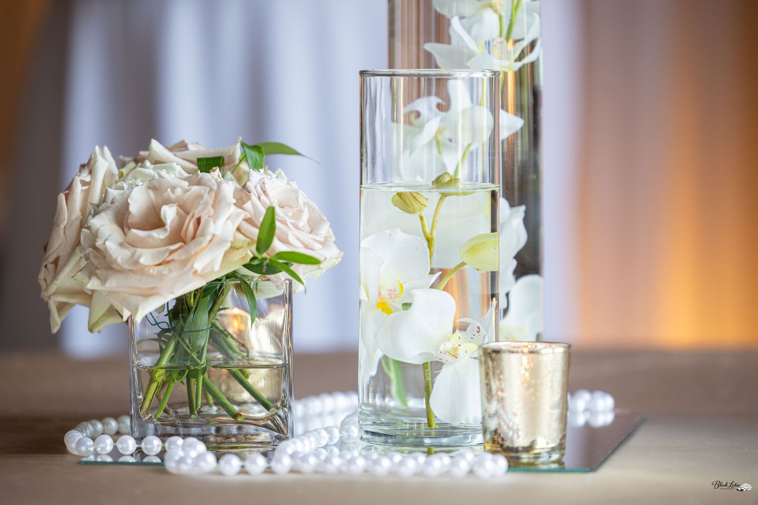 Elegant wedding centerpiece with blush roses, white orchids in glass vases, pearl strand, and gold candle on mirrored surface.