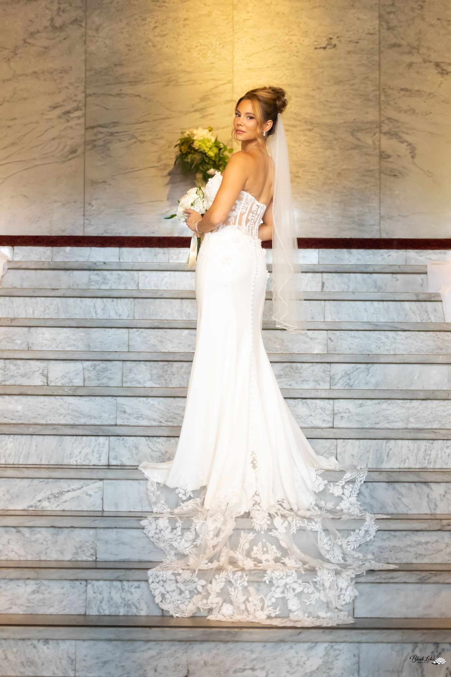 Bride in a white lace mermaid gown with train and veil holds bouquet on marble stairs.