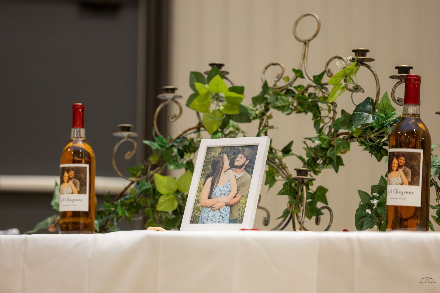 Wedding table display with framed couple photo, ivy garlands, candelabras, and two wine bottles on white linen.