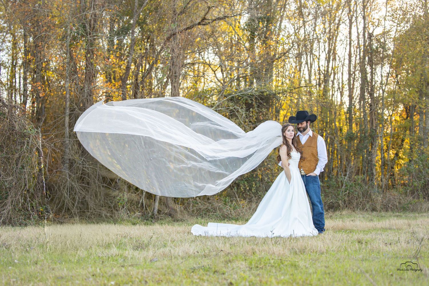 Bride and groom kiss outdoors as wedding veil flows dramatically in the wind with autumn trees in background.