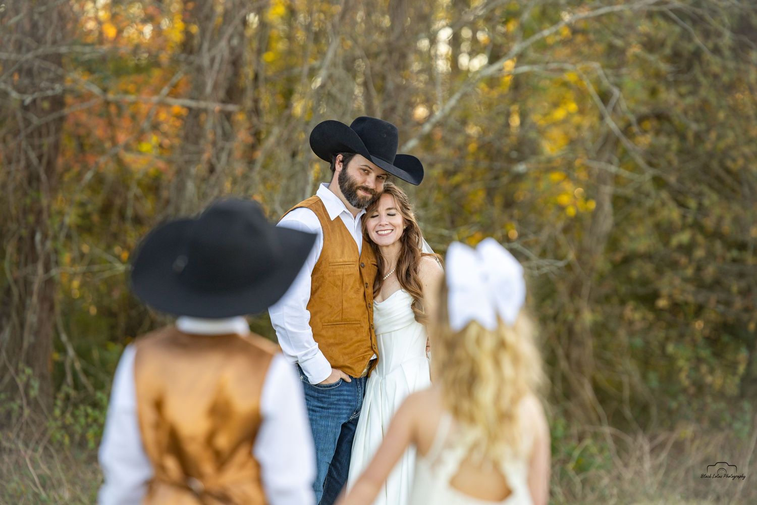 Rustic country wedding couple in black cowboy hats embrace outdoors, with guests in foreground amid autumn trees.