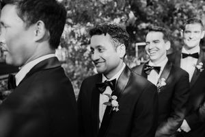 Smiling groom with boutonniere surrounded by groomsmen in tuxedos in a black and white outdoor wedding photo.