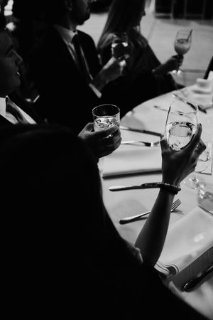 Black and white photo of wedding guests clinking glasses at elegantly set reception dinner table.