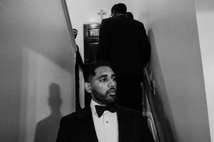 Black and white photo of groomsman in tuxedo standing in stairway at wedding venue.