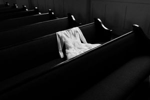 A white wedding dress draped over dark wooden church pews in a black and white photograph.