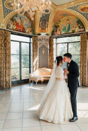 Bride and groom share romantic moment in ornate ballroom with gilded arched murals and chandelier.