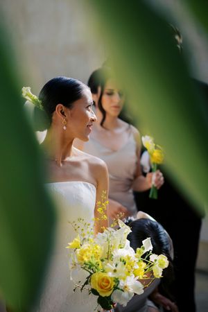 Bride in strapless white gown holding yellow and white bouquet stands with bridesmaid in pink dress during wedding ceremony.