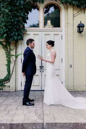 Bride reads vows to groom outside yellow building with white doors and ivy on wedding day.