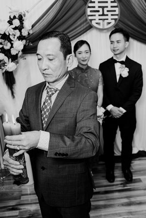 Black and white photo of a man in a suit lighting candles at a Chinese wedding ceremony with bride and groom in background.