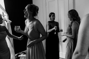 Black and white photo of bridesmaids in elegant gowns laughing and holding drinks during wedding preparations.