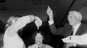 Black and white photo of three people at an event, two raising hands while a woman stands between them.