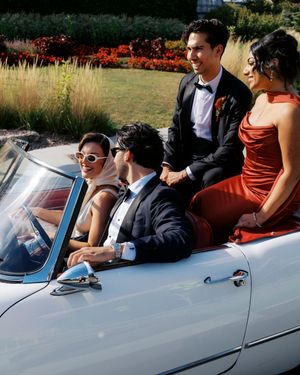 Wedding party riding in a white convertible classic car, dressed in formal attire with a red bridesmaid gown.
