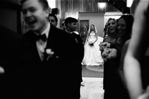 Black and white wedding photo of bride in white gown standing in doorway surrounded by groom and guests.