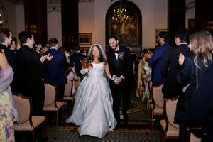 Bride and groom walk down the aisle at elegant indoor wedding ceremony while guests applaud in formal attire.