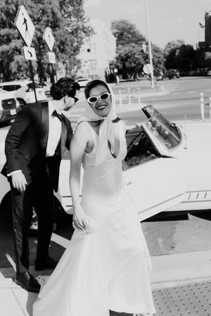 Black and white photo of a smiling bride in sunglasses and white gown beside a vintage convertible car.