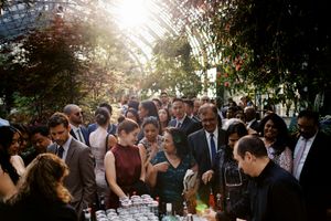 Elegant indoor garden party with diverse guests mingling under a sunlit glass greenhouse canopy.
