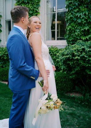 Bride in white gown holding bouquet laughs with groom in blue suit outdoors near ivy-covered building.