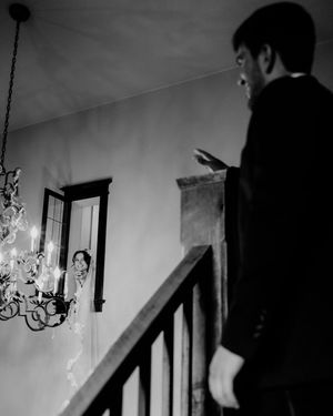 Black and white wedding photo of groom on staircase gesturing toward bride standing in doorway with chandelier.