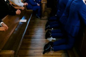 People in blue suits sitting in church pews during a wedding ceremony, showing clasped hands and dress shoes.