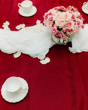 Elegant red tablecloth with white teacups, rose petals, and a pink rose bouquet centerpiece for a romantic wedding setting.
