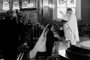 Black and white wedding ceremony photo of couple kneeling before priest in elegant church.
